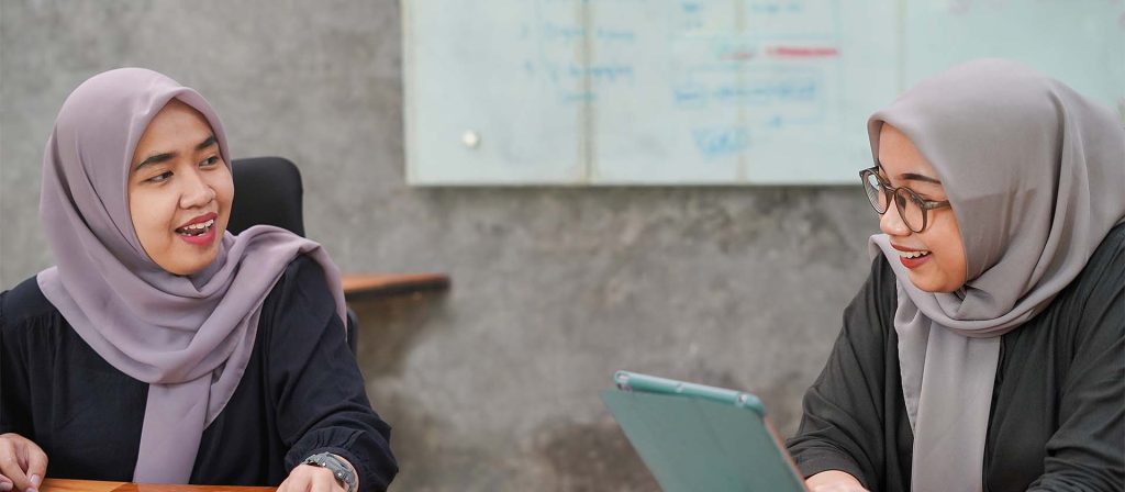 Two Asian women wearing hijabs talking to each other in office.