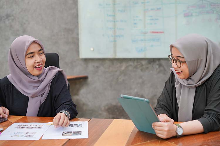 Two Asian women wearing hijabs talking to each other in office.