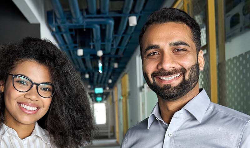 A woman and man stand side-by-side in an office corridor, smiling at the camera